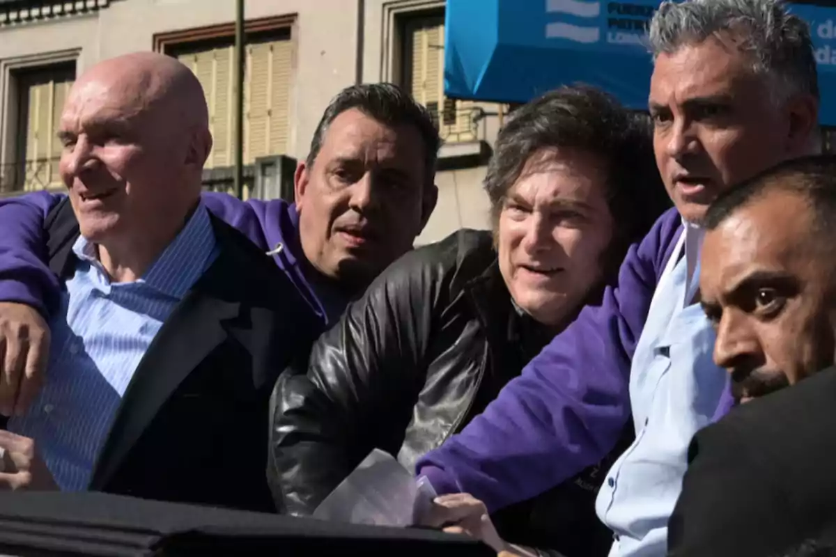 Cinco hombres abrazados en la calle durante un evento al aire libre con edificios y un toldo azul de fondo Cinco hombres abrazados en la calle durante un evento al aire libre con edificios y un toldo azul de fondo