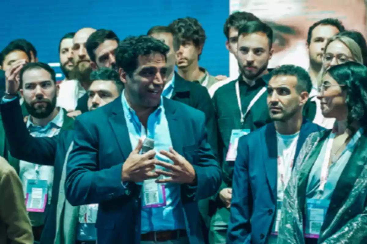 A group of people dressed formally is gathered at an event; one of them is holding a microphone and appears to be speaking while the others listen alertly.