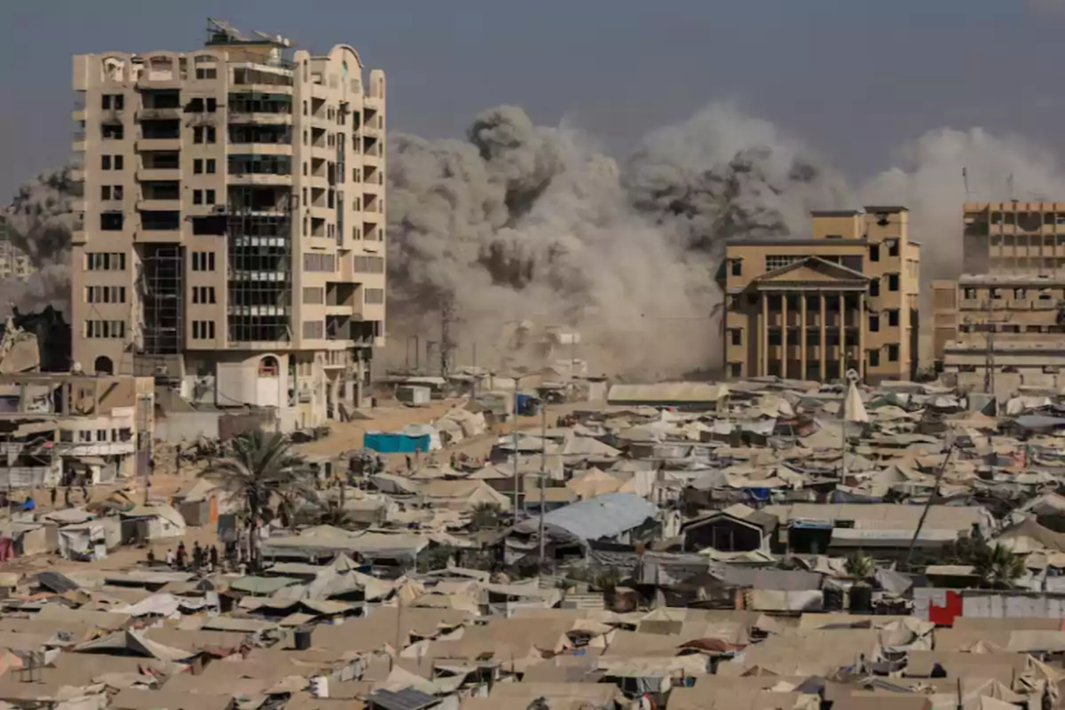 Partially damaged tall buildings and a large cloud of smoke in the background, while in the foreground numerous makeshift tents and people in a camp can be seen. Partially damaged tall buildings and a large cloud of smoke in the background, while in the foreground numerous makeshift tents and people in a camp can be seen.