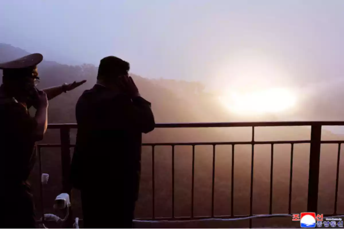 Two people with their backs turned watch a bright explosion in the distance from a lookout with a railing.