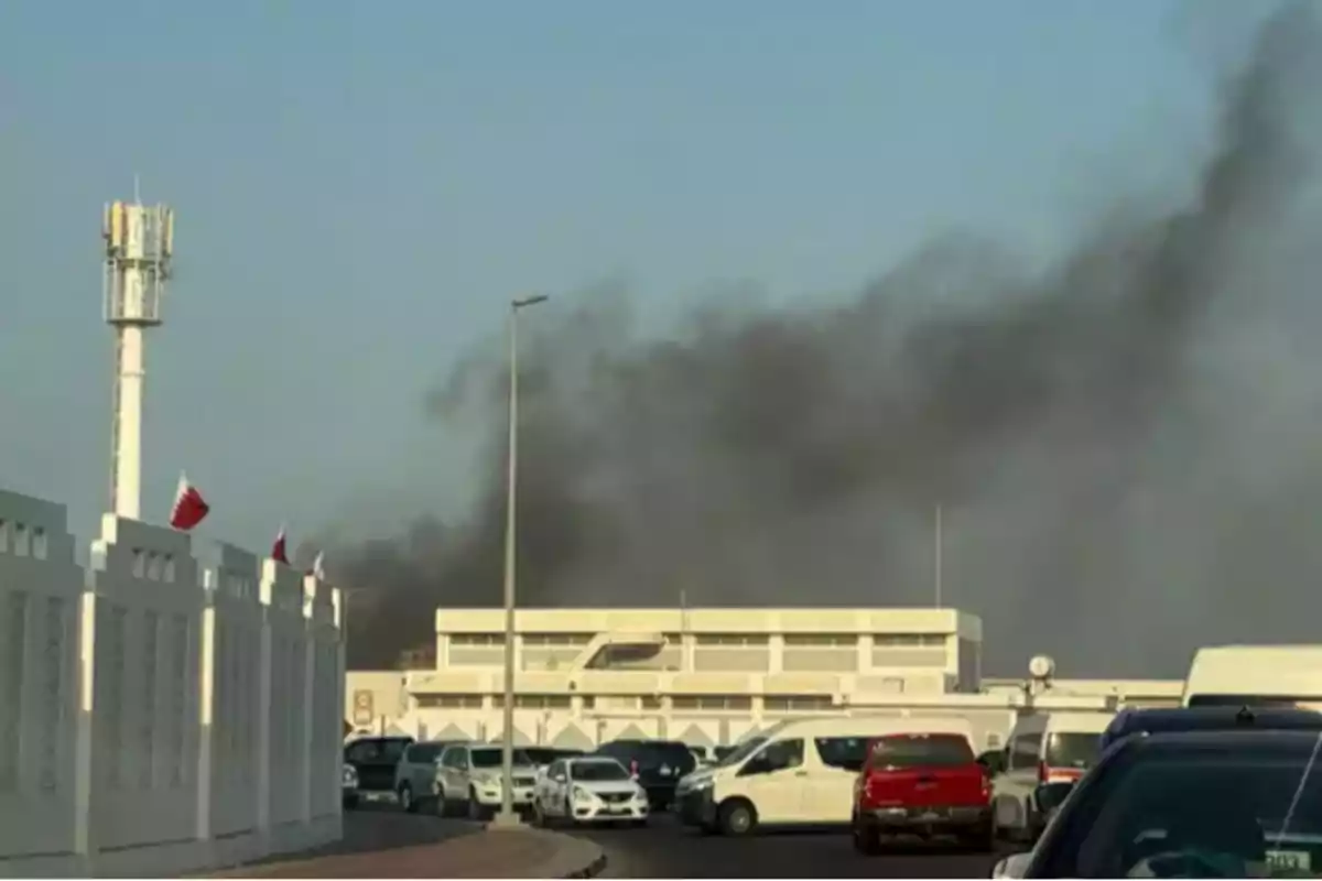 Humo negro saliendo de un edificio blanco mientras varios autos están estacionados en la calle bajo un cielo despejado
