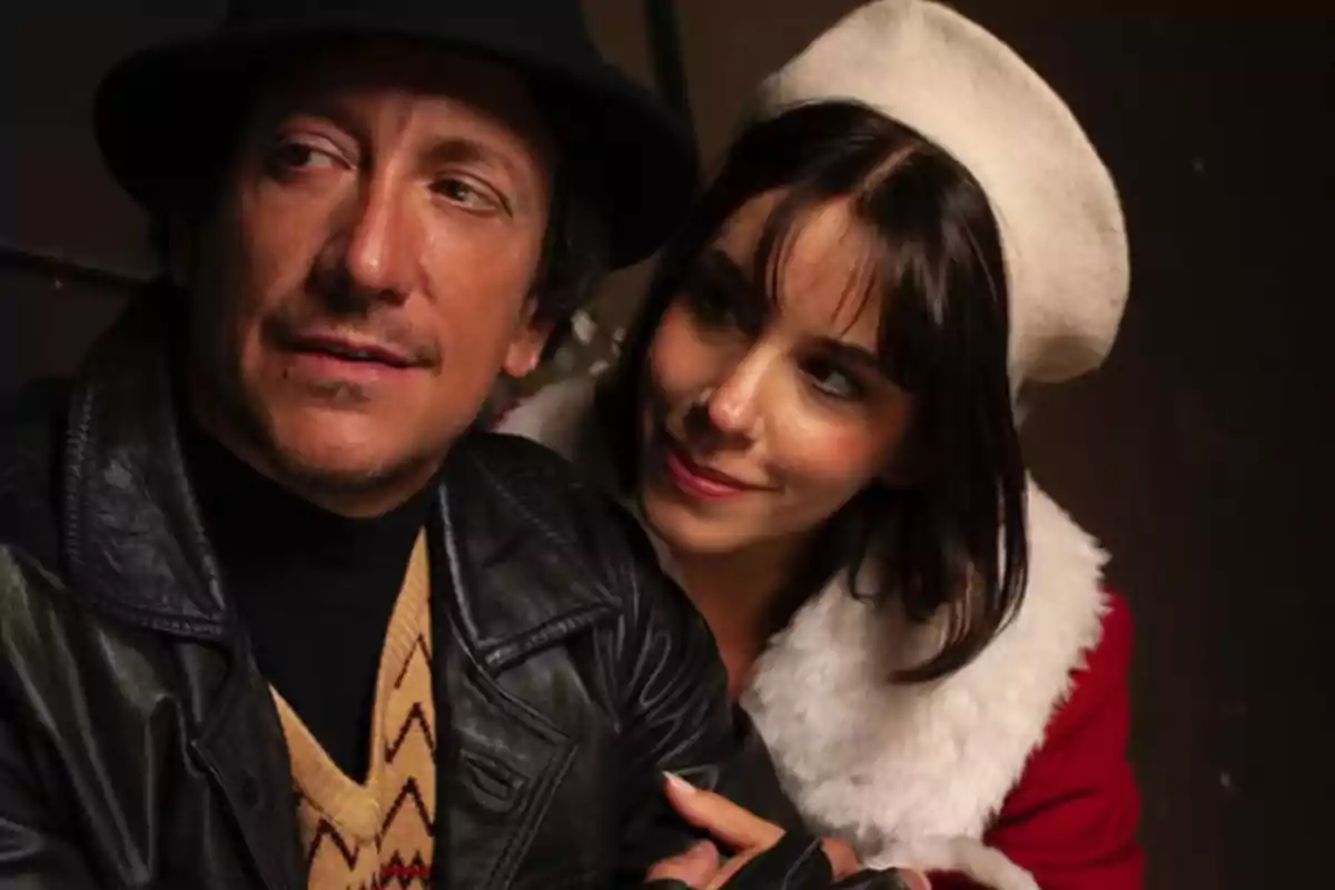 A man wearing a black hat and a leather jacket is accompanied by a woman wearing a white beret and a red coat with white faux fur details; she looks at him with an affectionate expression. A man wearing a black hat and a leather jacket is accompanied by a woman wearing a white beret and a red coat with white faux fur details; she looks at him with an affectionate expression.