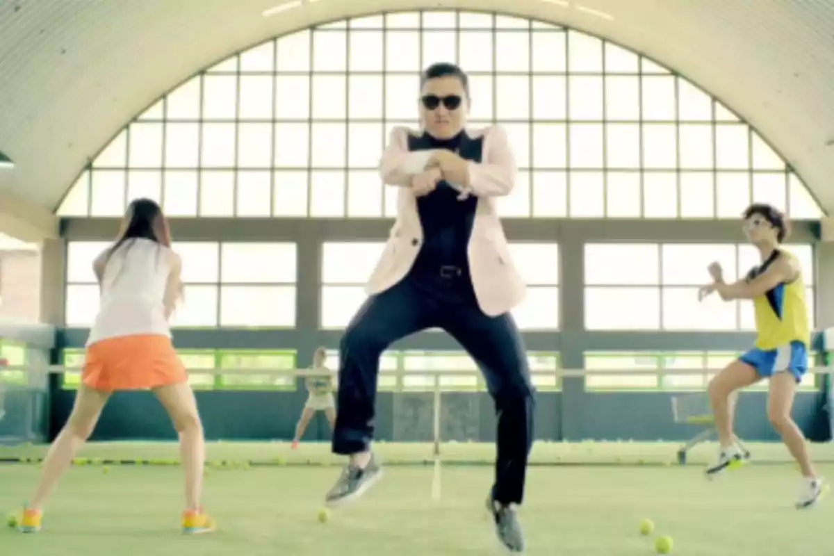 Persona con gafas de sol y chaqueta clara bailando en una cancha de tenis cubierta acompañado de otras personas