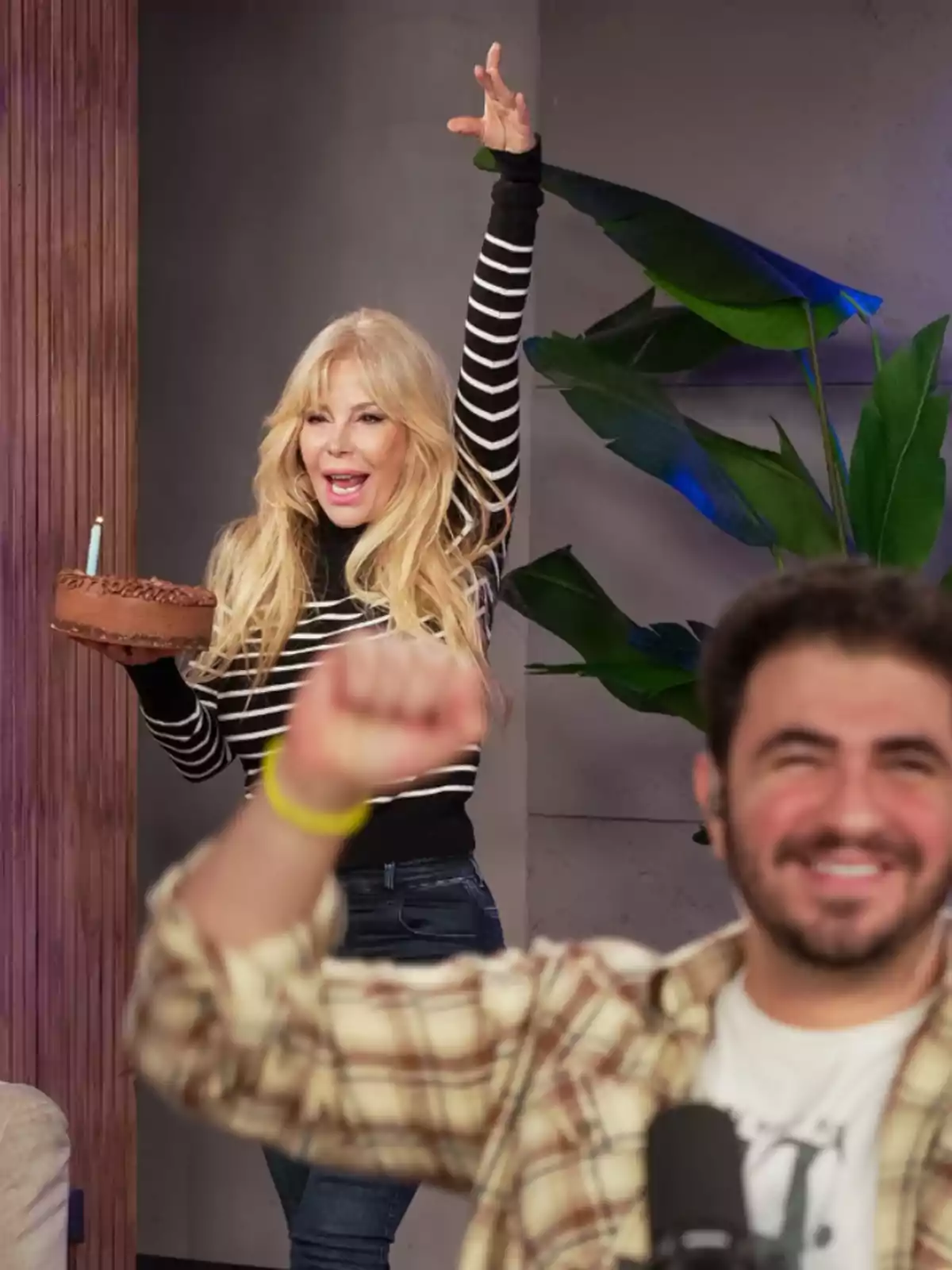 Smiling blonde woman in a striped sweater holds a chocolate cake with a candle while raising one arm, and a man in front smiles with his fist in the air. Smiling blonde woman in a striped sweater holds a chocolate cake with a candle while raising one arm, and a man in front smiles with his fist in the air.