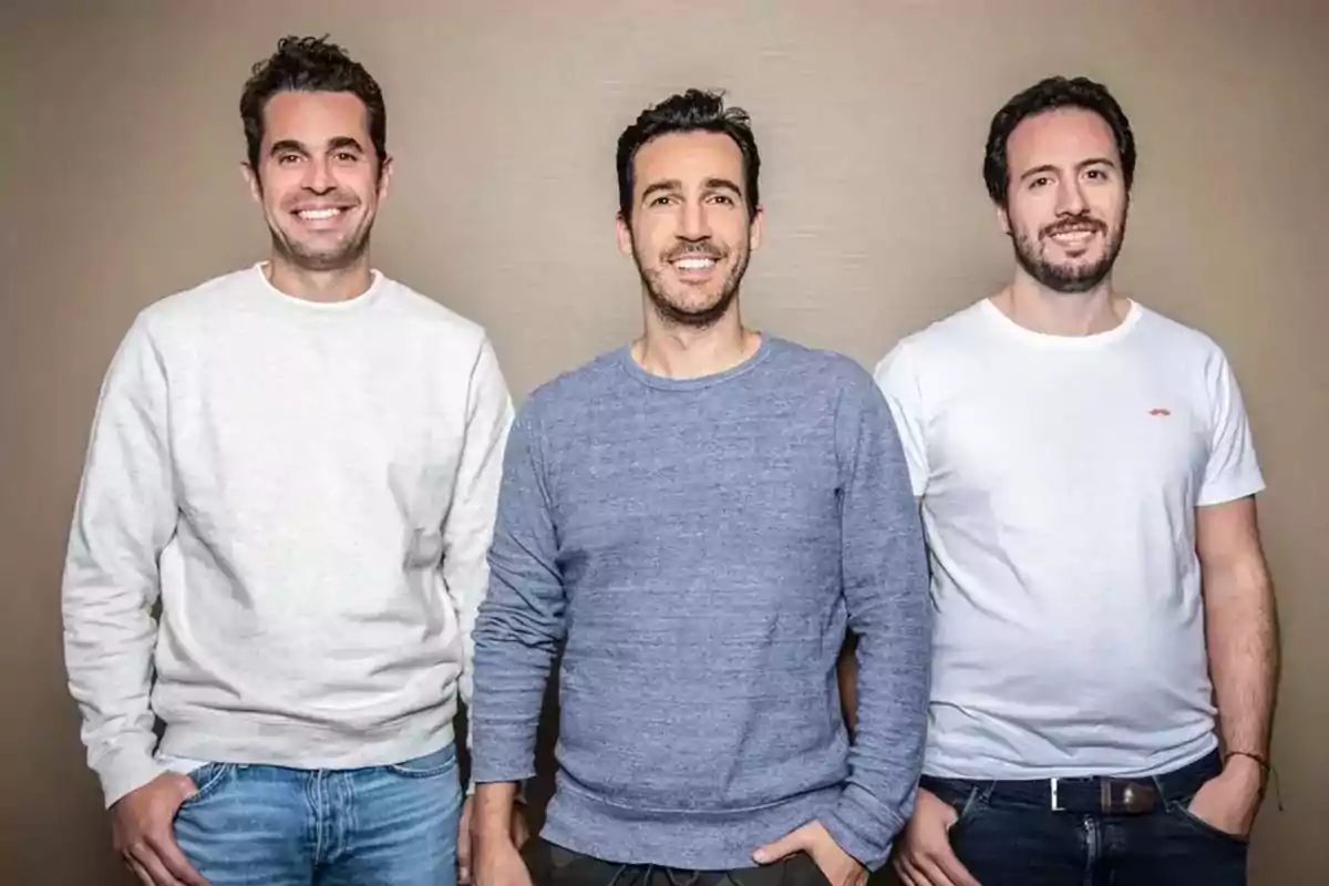 Three young men pose smiling in front of a beige wall, each wearing casual clothes. Three young men pose smiling in front of a beige wall, each wearing casual clothes.