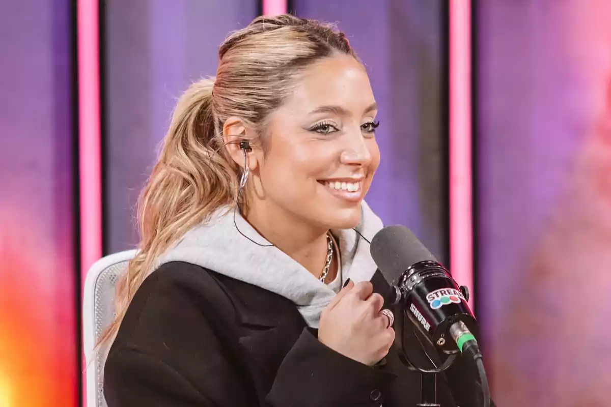 Young woman smiling while speaking in front of a microphone in a recording studio with colorful lights in the background Young woman smiling while speaking in front of a microphone in a recording studio with colorful lights in the background