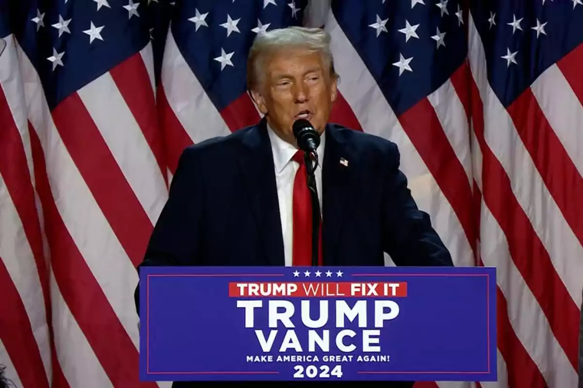 Hombre de traje oscuro y corbata roja hablando en un podio con letrero de campaña que dice Trump Vance 2024 frente a varias banderas de Estados Unidos