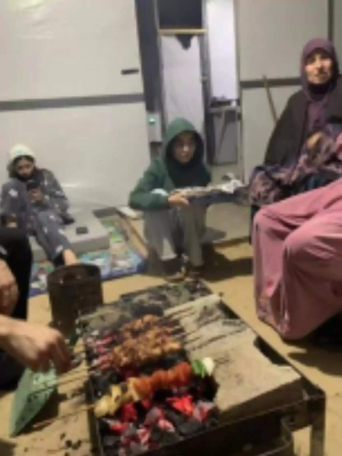 Several people sitting around a grill roasting skewers in a simple indoor setting Several people sitting around a grill roasting skewers in a simple indoor setting