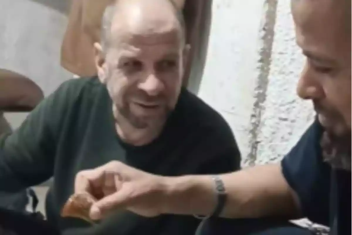 Two men sitting on the floor share food while one of them smiles.