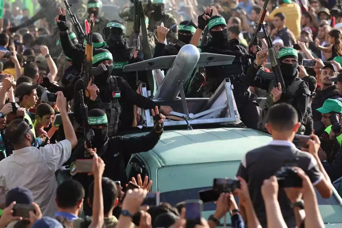 Armed militiamen in black uniforms and green bandanas surround a vehicle carrying a drone while a crowd watches and takes photos.