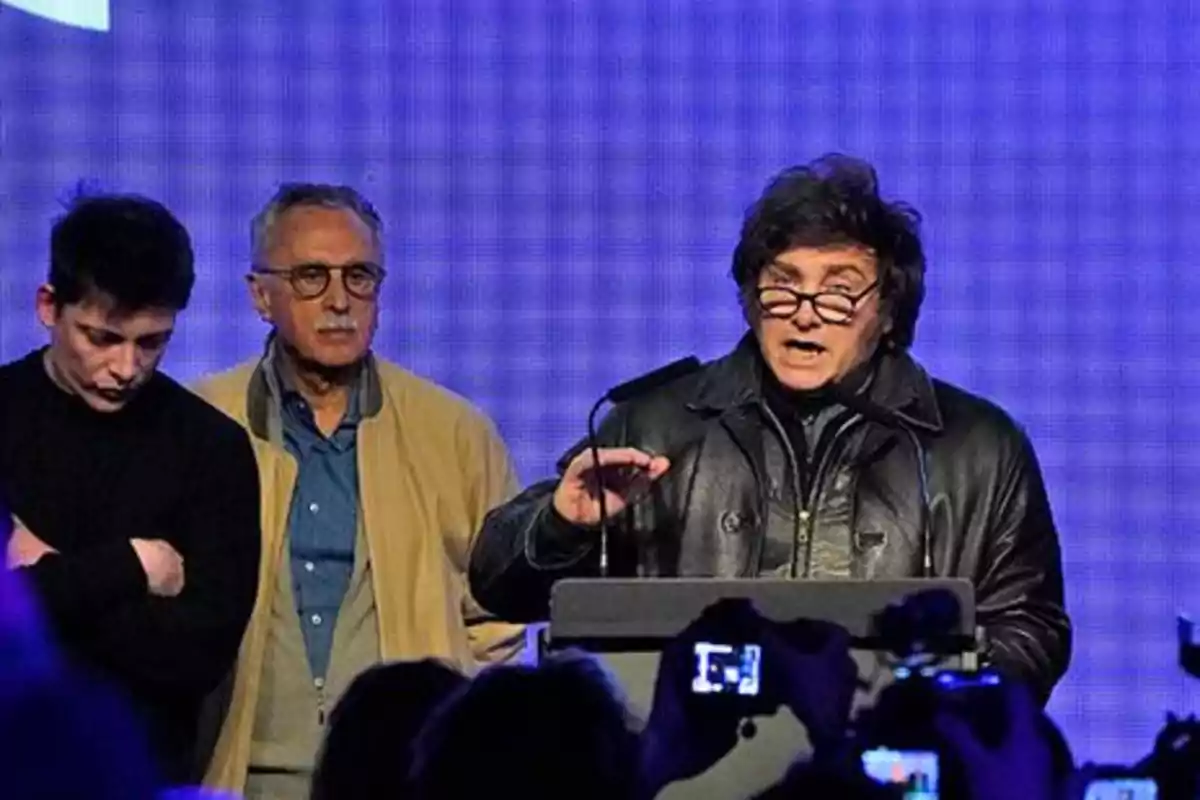 A man in a leather jacket and glasses speaks at a podium in front of an audience while two people accompany him on stage. A man in a leather jacket and glasses speaks at a podium in front of an audience while two people accompany him on stage.