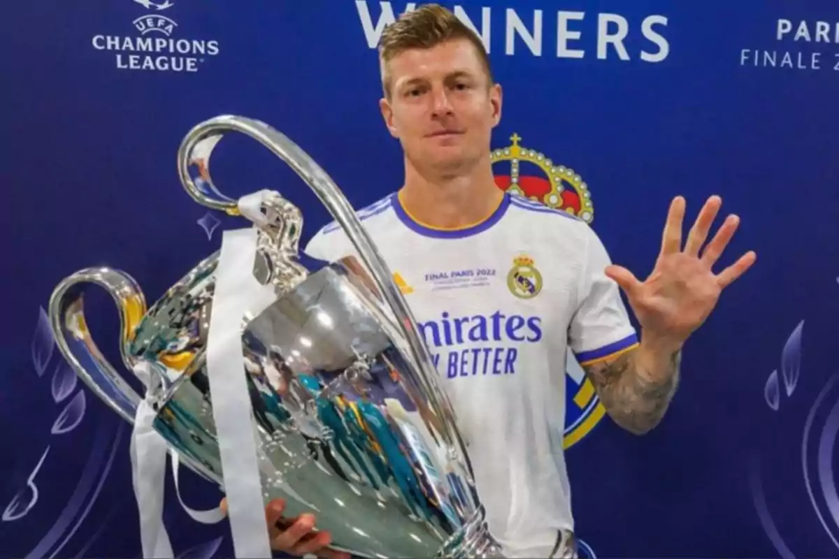 Futbolista del Real Madrid sosteniendo el trofeo de la Champions League y mostrando cinco dedos frente a un fondo azul con el logo del torneo. Futbolista del Real Madrid sosteniendo el trofeo de la Champions League y mostrando cinco dedos frente a un fondo azul con el logo del torneo.