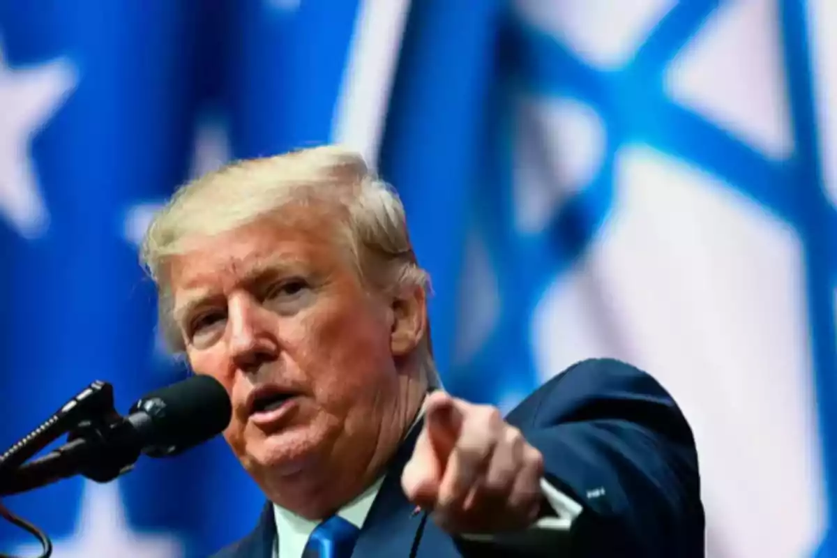 Blond-haired man speaking into a microphone and pointing forward with blurred flags in the background