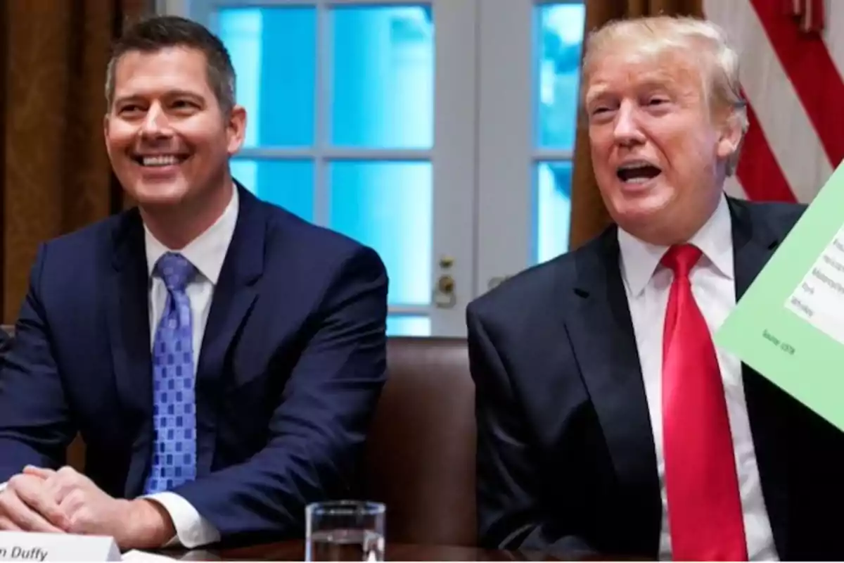 Two men in suits sitting at a table, one of them holding a green sheet while they smile and talk in a room with windows in the background.