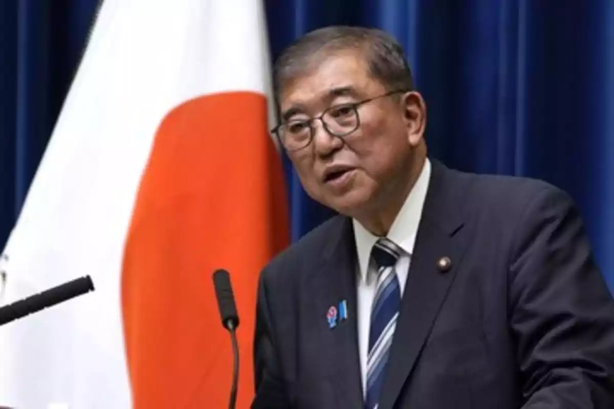 Man with glasses and a dark suit speaking in front of a microphone with a Japanese flag in the background