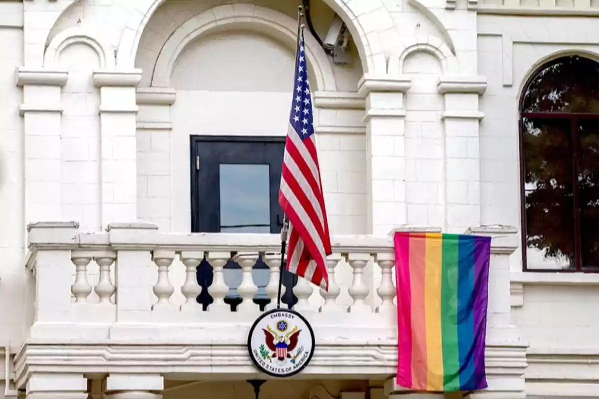Fachada de un edificio blanco con una bandera de Estados Unidos y una bandera arcoíris colgadas en el balcón junto a un escudo que dice United States of America Fachada de un edificio blanco con una bandera de Estados Unidos y una bandera arcoíris colgadas en el balcón junto a un escudo que dice United States of America