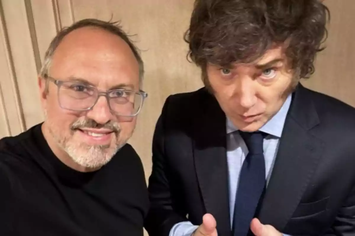 Two men pose together for a selfie; one of them is wearing glasses and a black t-jersey, while the other is dressed in a dark suit and blue tie.