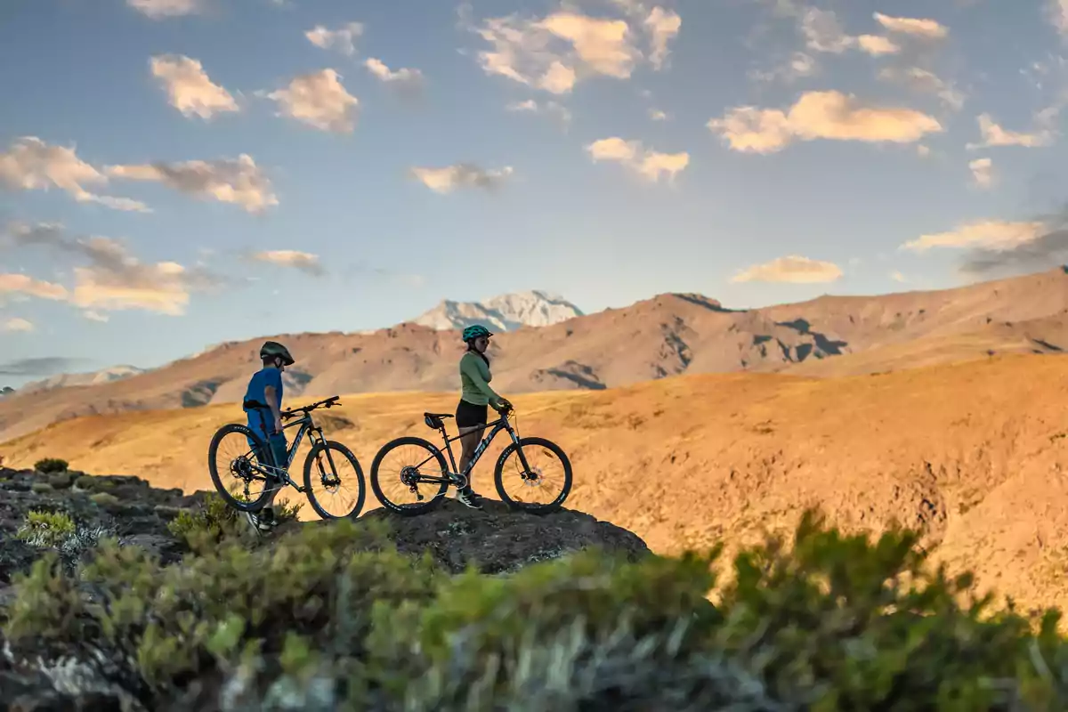 Dos personas con bicicletas de montaña disfrutan de una vista panorámica en un paisaje árido con montañas al fondo y cielo parcialmente nublado Dos personas con bicicletas de montaña disfrutan de una vista panorámica en un paisaje árido con montañas al fondo y cielo parcialmente nublado