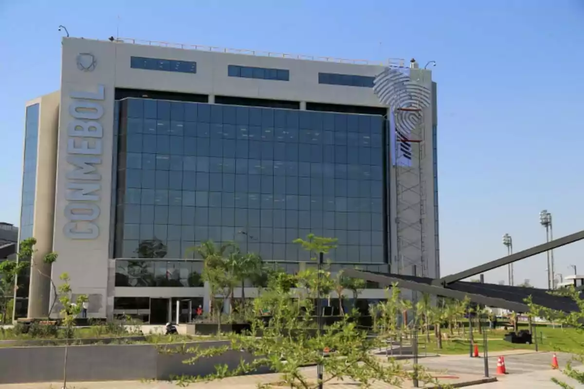 Edificio moderno de la sede de la CONMEBOL con fachada de vidrio y vegetación en el exterior en un día soleado