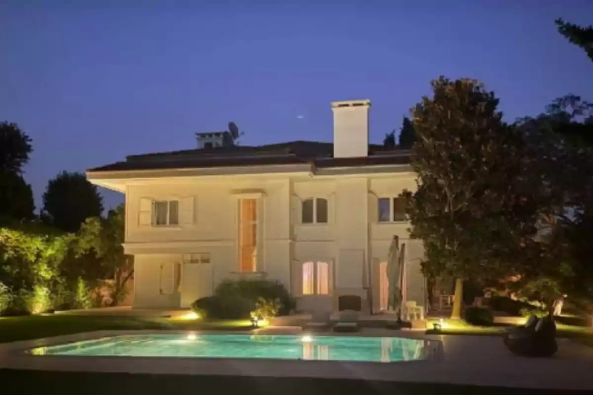 Casa grande de dos pisos con piscina iluminada en el jardín durante el atardecer rodeada de árboles y vegetación Casa grande de dos pisos con piscina iluminada en el jardín durante el atardecer rodeada de árboles y vegetación