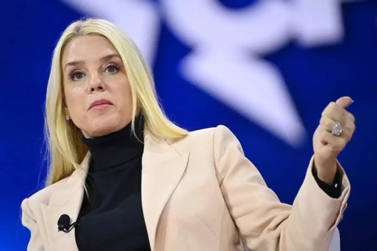 Mujer rubia con chaqueta clara y suéter negro hablando en un escenario con fondo azul Mujer rubia con chaqueta clara y suéter negro hablando en un escenario con fondo azul