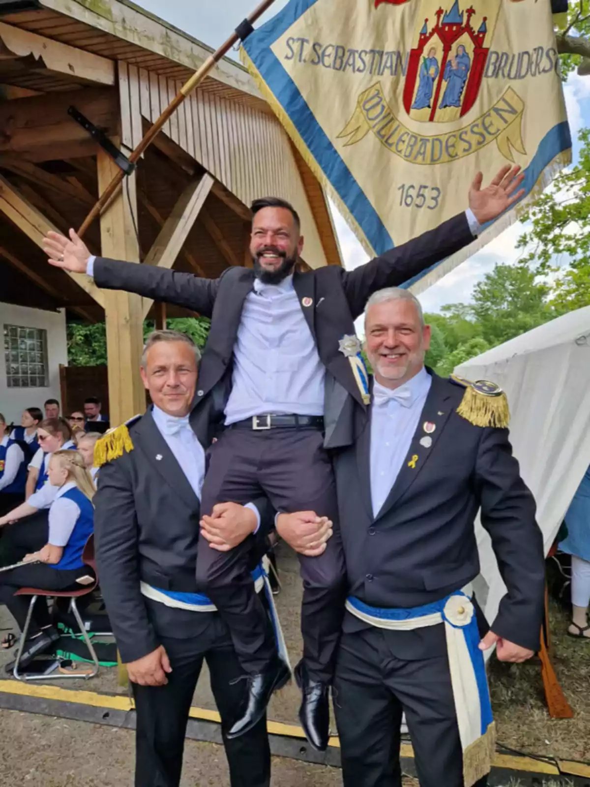 Tres hombres vestidos con trajes formales y bandas azules posan sonrientes, dos de ellos levantan al tercero mientras al fondo se observa una bandera y varias personas sentadas Tres hombres vestidos con trajes formales y bandas azules posan sonrientes, dos de ellos levantan al tercero mientras al fondo se observa una bandera y varias personas sentadas