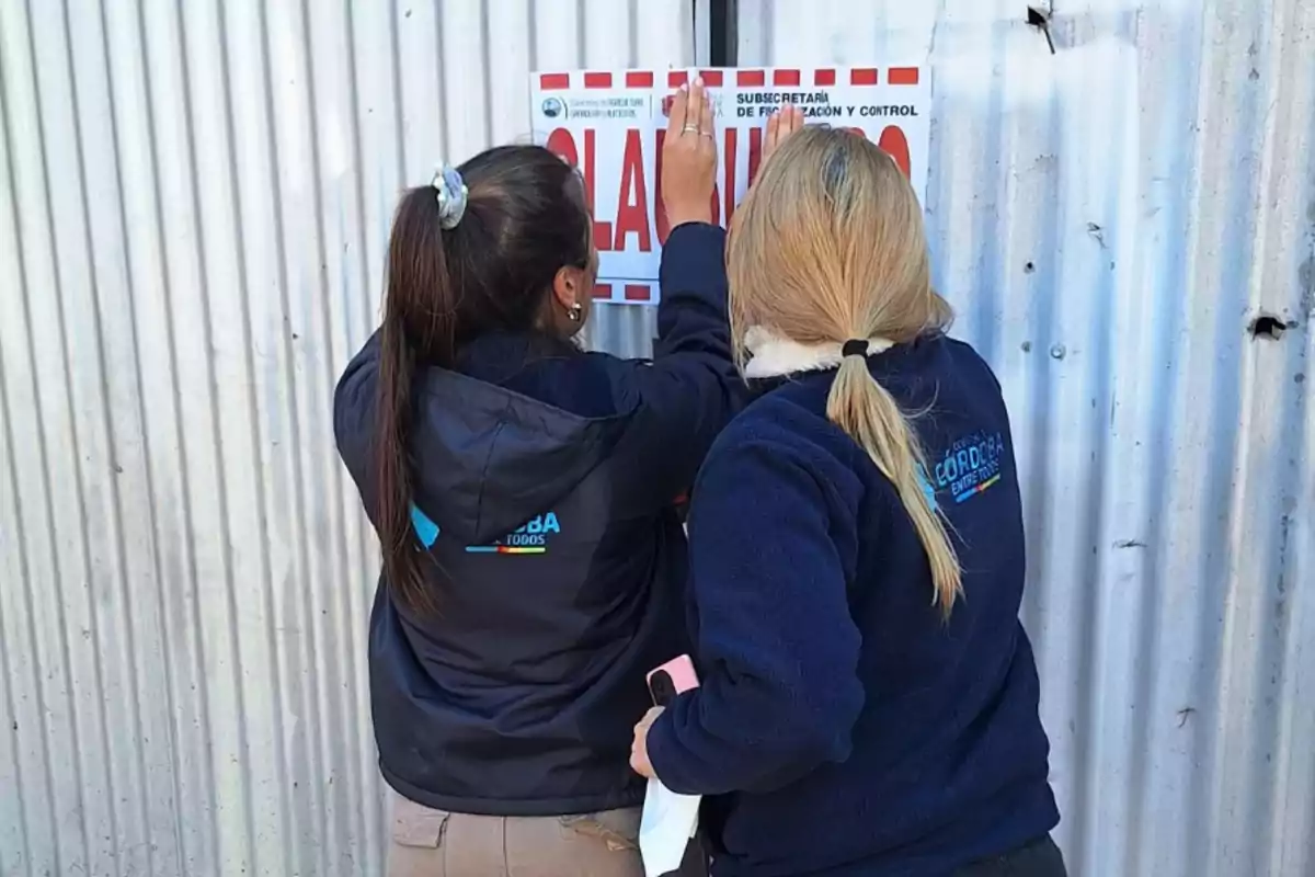 Dos mujeres de espaldas colocan un cartel de clausura en una pared de chapa mientras usan camperas con logos institucionales