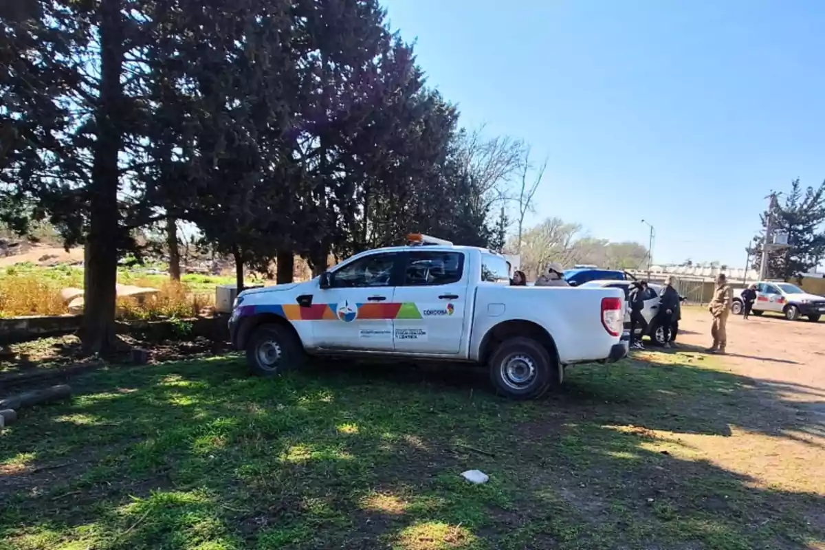 Camioneta blanca de protección civil estacionada en un área verde junto a varios árboles y personas al fondo