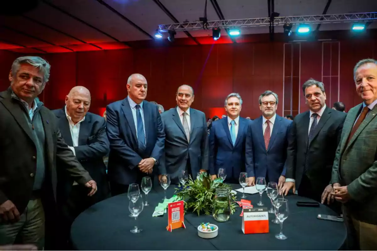Eight men dressed in suits and jackets pose around a round table at a formal event with glasses and decorations in the center, in a hall lit with blue and red lights.