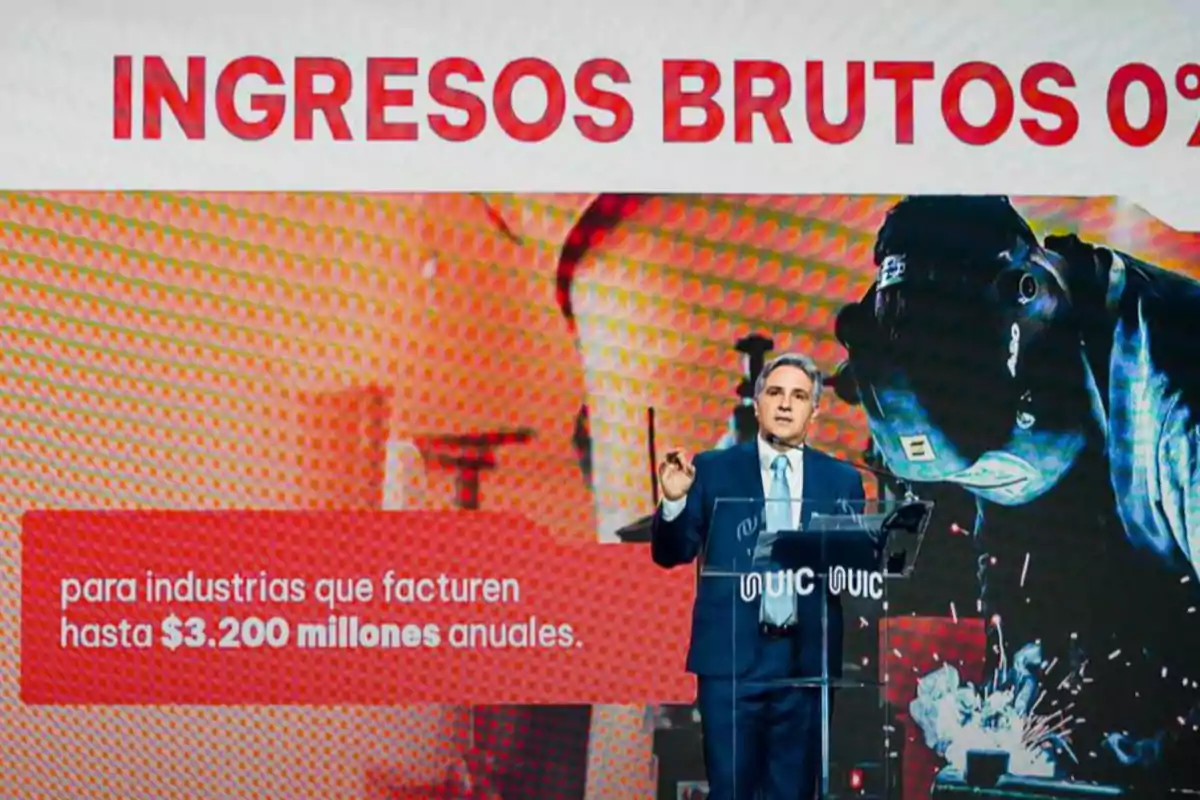 A man in a suit speaks on a stage in front of a screen displaying the text A man in a suit speaks on a stage in front of a screen displaying the text