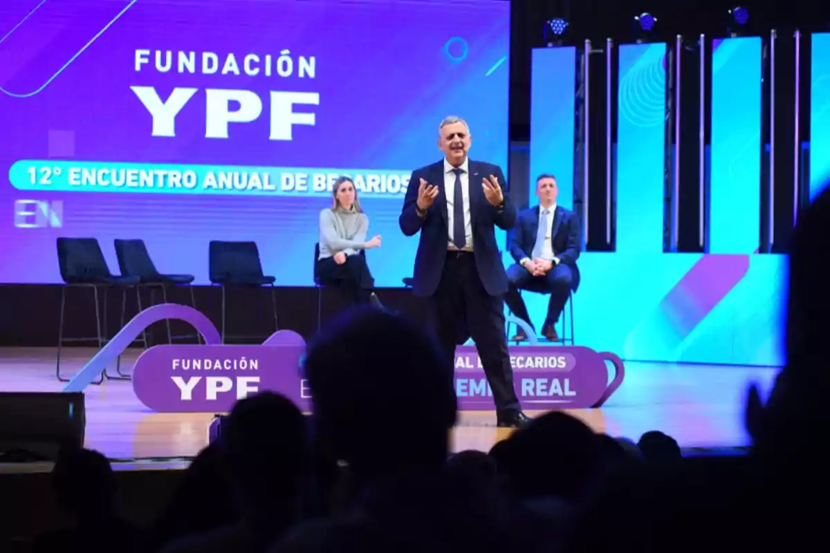 A man in a suit speaks on stage during a YPF Foundation event while two seated people watch him and the audience is in the foreground.