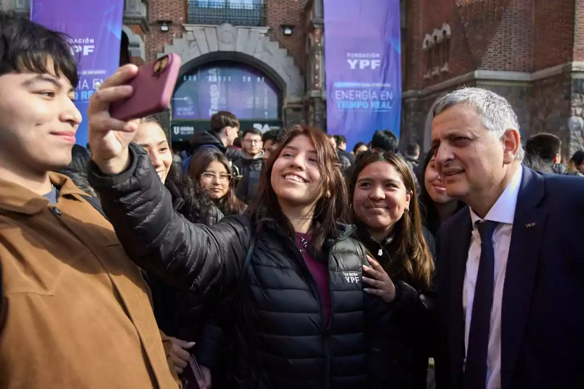 A group of young people smile as they take a selfie with a man in a suit in front of a building with YPF Foundation signs at an outdoor event. A group of young people smile as they take a selfie with a man in a suit in front of a building with YPF Foundation signs at an outdoor event.