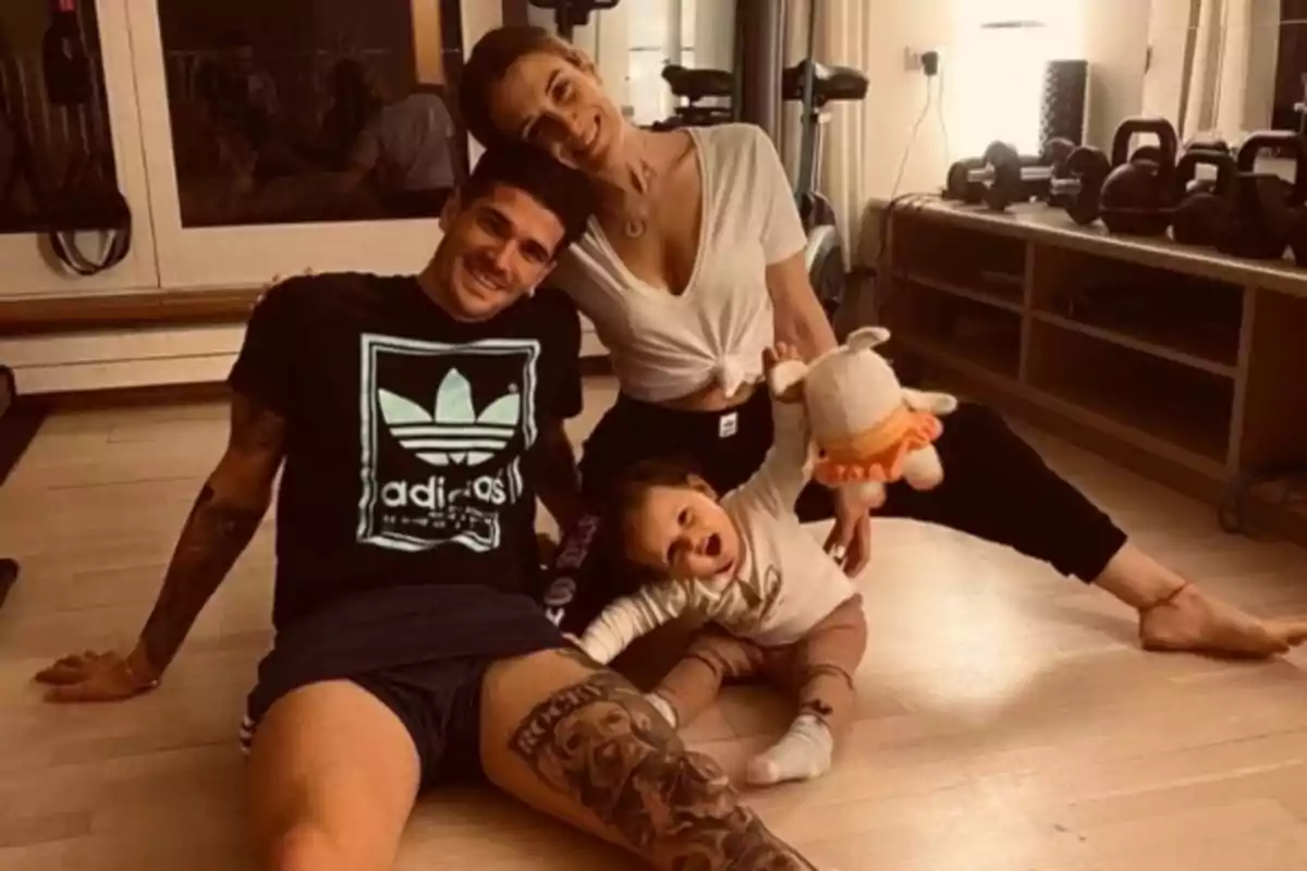 A smiling family sits on the floor of a room with weights and exercise equipment in the background; the mother and father are hugging their young daughter, who is holding a stuffed animal. A smiling family sits on the floor of a room with weights and exercise equipment in the background; the mother and father are hugging their young daughter, who is holding a stuffed animal.