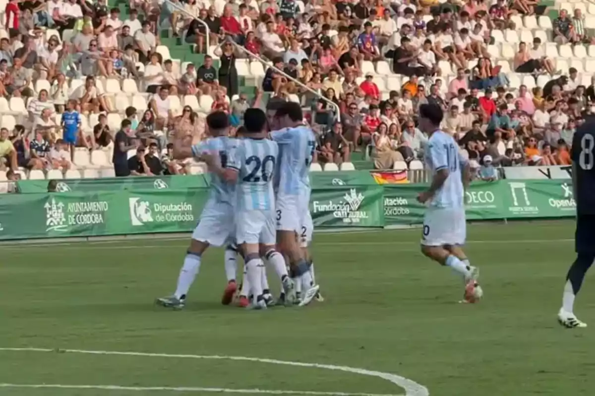 Soccer players in light blue and white uniforms celebrate a goal on the field while the crowd watches from the stands. Soccer players in light blue and white uniforms celebrate a goal on the field while the crowd watches from the stands.