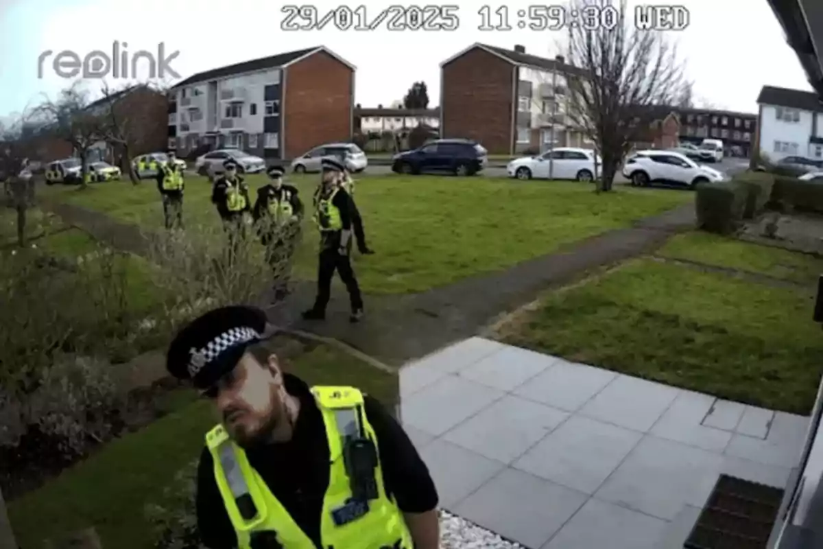 Several police officers wearing yellow vests walk through a residential garden while one of them approaches the security camera. Several police officers wearing yellow vests walk through a residential garden while one of them approaches the security camera.