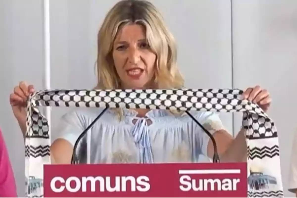 Blonde woman holding a black and white keffiyeh while speaking at a podium with the Comuns Sumar logo
