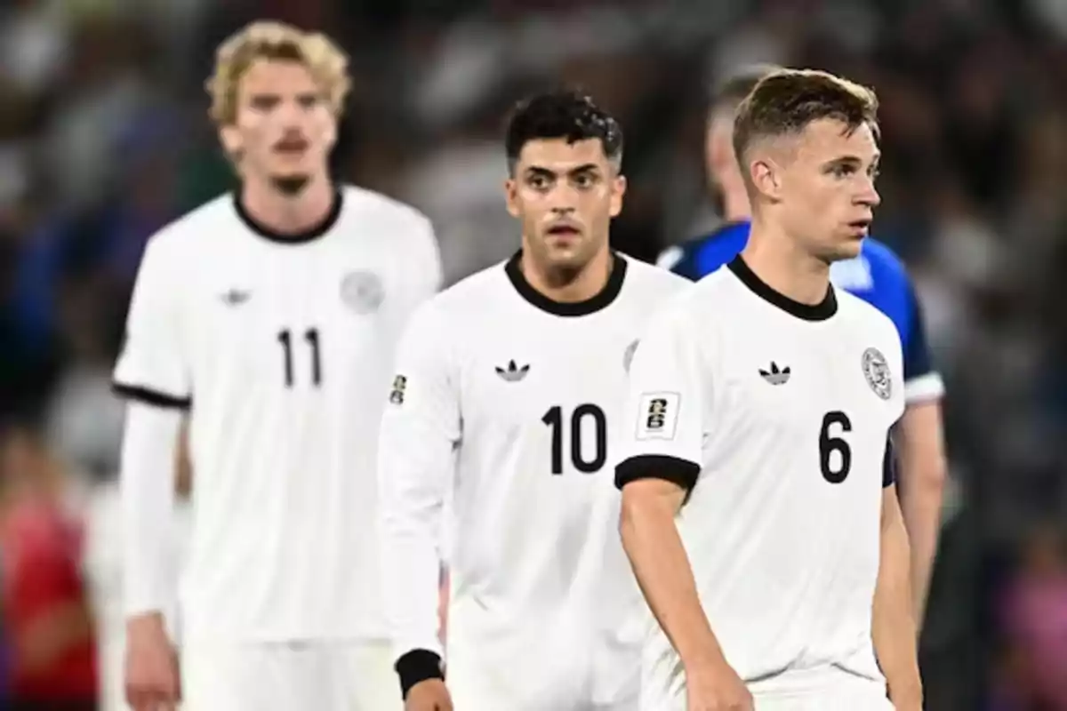Tres futbolistas con uniforme blanco caminan en el campo con expresiones serias durante un partido Tres futbolistas con uniforme blanco caminan en el campo con expresiones serias durante un partido