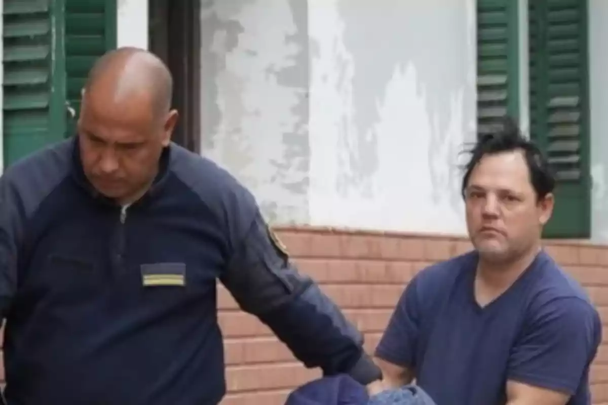 Un policía escolta a un hombre de cabello oscuro y camiseta azul frente a una pared blanca con ventanas verdes