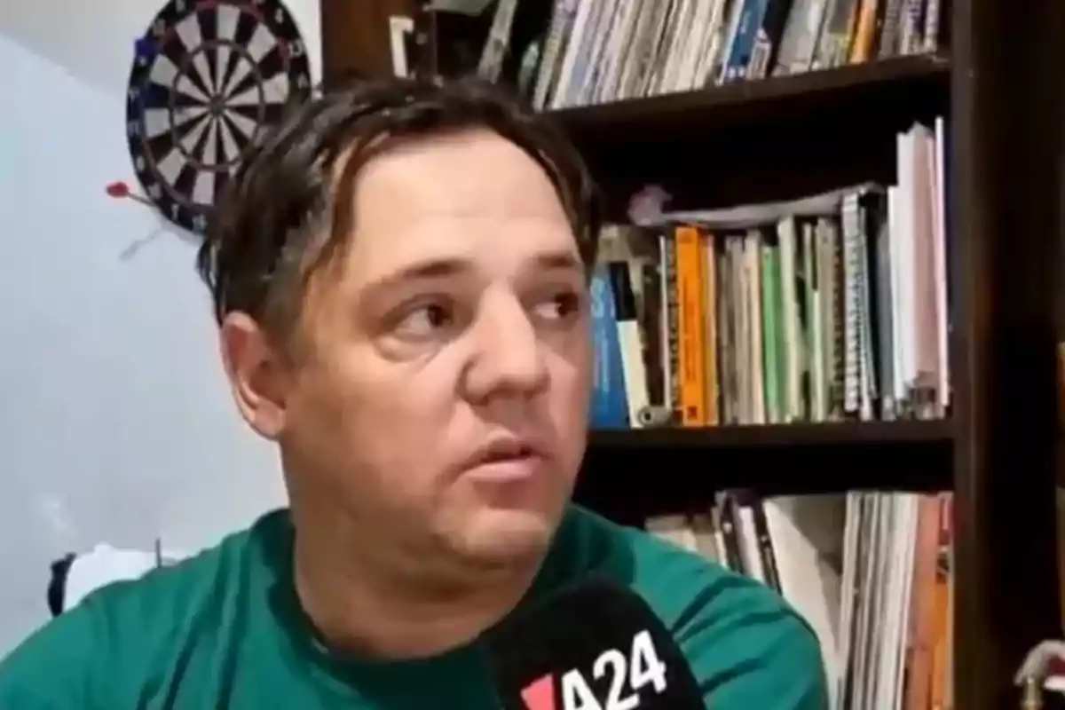Hombre con camiseta verde siendo entrevistado por un canal de noticias frente a una estantería con libros y un tablero de dardos en la pared. Hombre con camiseta verde siendo entrevistado por un canal de noticias frente a una estantería con libros y un tablero de dardos en la pared.