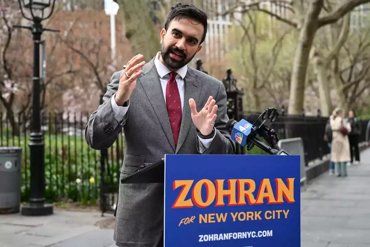 Un hombre con barba y traje gris da un discurso al aire libre frente a un podio azul con el nombre Zohran y varios micrófonos de prensa Un hombre con barba y traje gris da un discurso al aire libre frente a un podio azul con el nombre Zohran y varios micrófonos de prensa