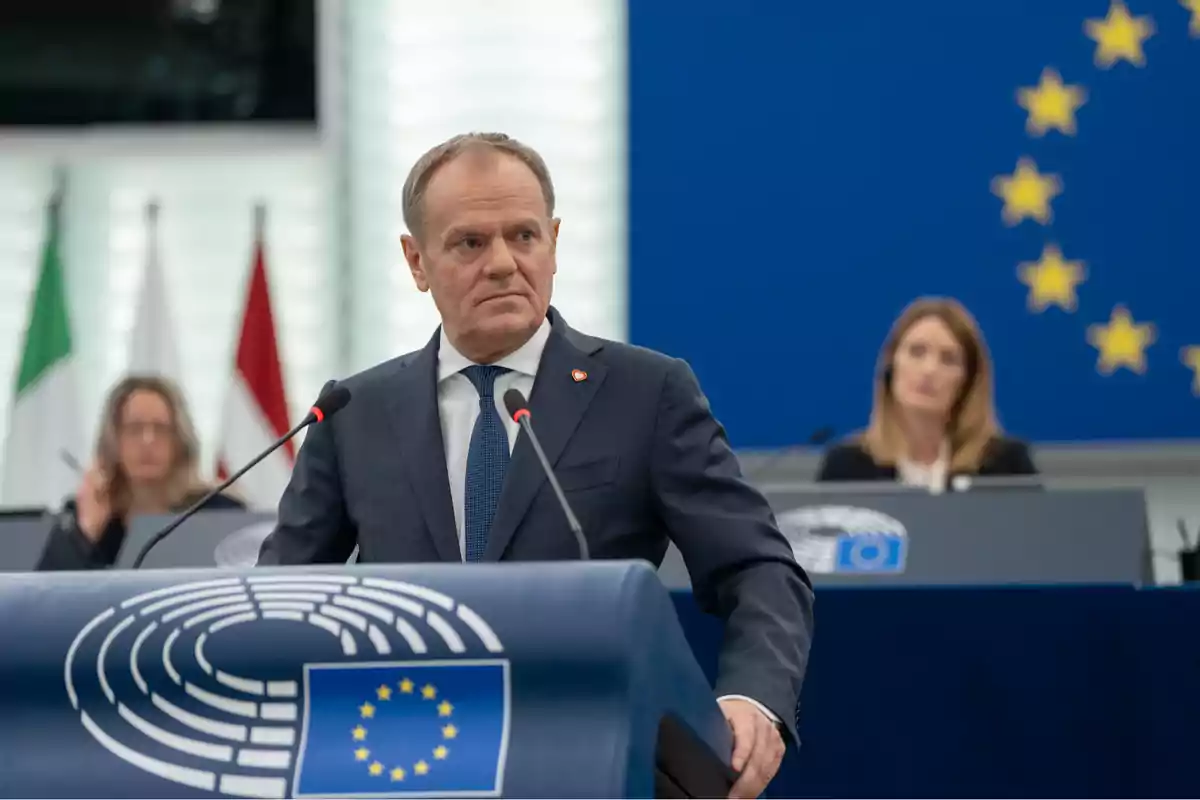 Un hombre de traje hablando en un podio con el emblema de la Unión Europea en un entorno de conferencia con banderas y personas al fondo.