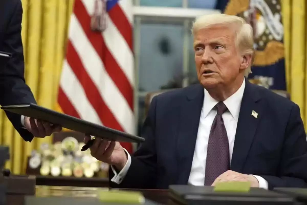 A man in a suit sitting at a desk receives a document from another person in an office with flags in the background. A man in a suit sitting at a desk receives a document from another person in an office with flags in the background.