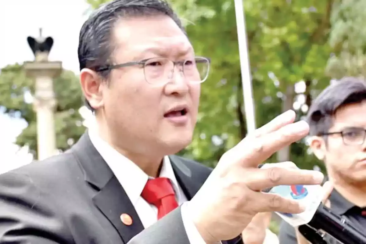 A man with glasses and a dark suit speaks while holding a microphone, with trees in the background.