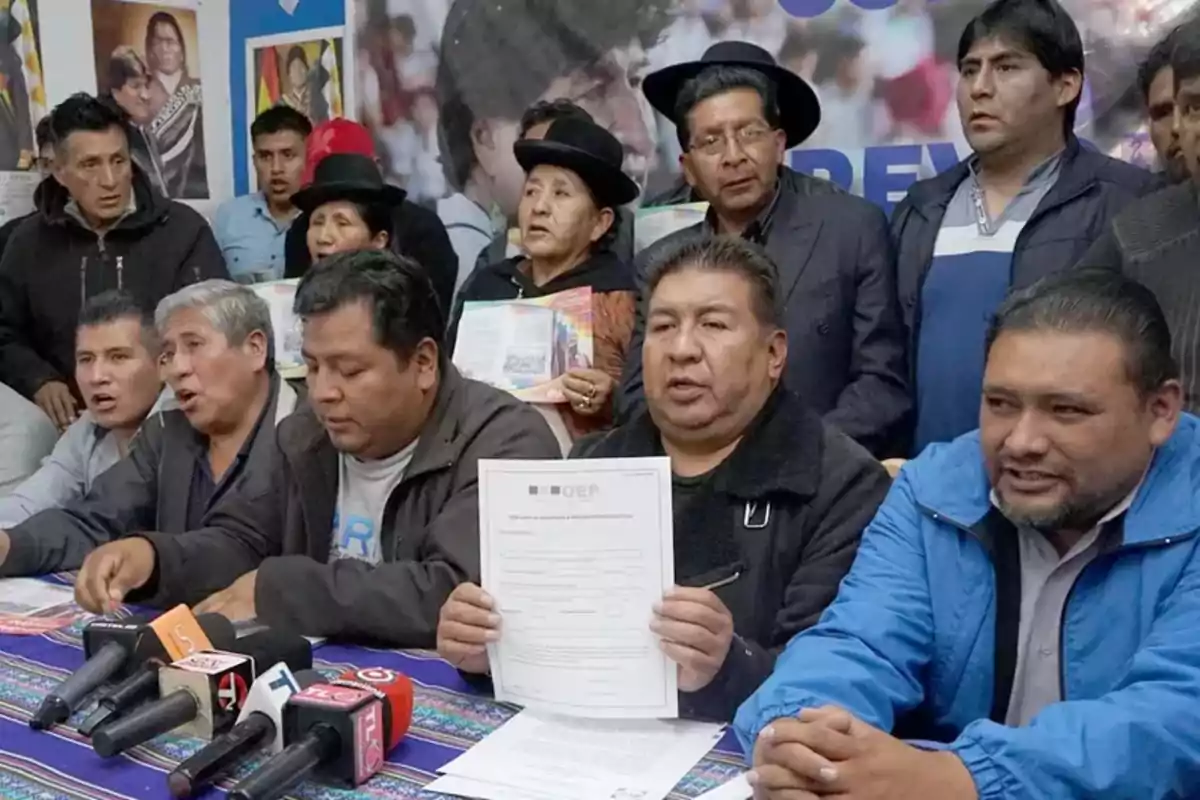 Un grupo de personas en una conferencia de prensa con micrófonos y documentos sobre una mesa.