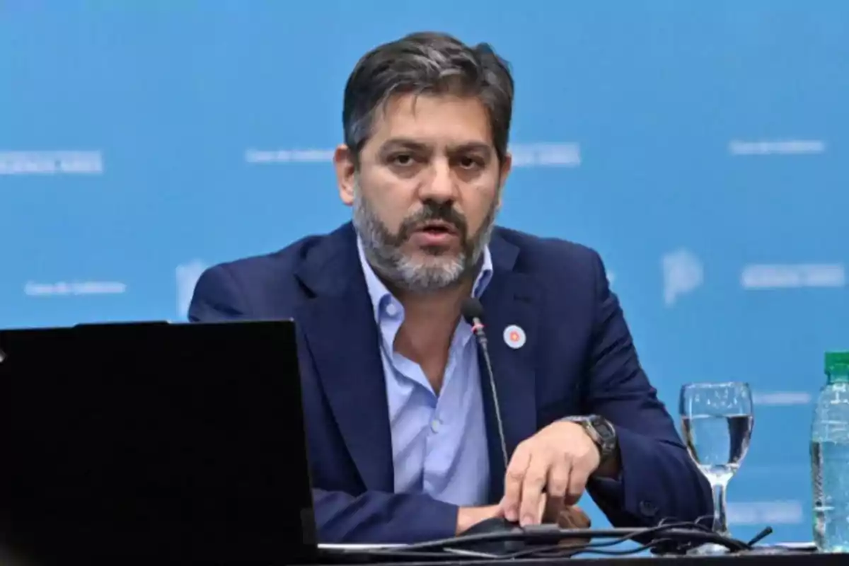 Un hombre con barba y traje oscuro habla en una conferencia de prensa frente a un micrófono, con una computadora portátil y un vaso de agua sobre la mesa, y un fondo azul claro. Un hombre con barba y traje oscuro habla en una conferencia de prensa frente a un micrófono, con una computadora portátil y un vaso de agua sobre la mesa, y un fondo azul claro.