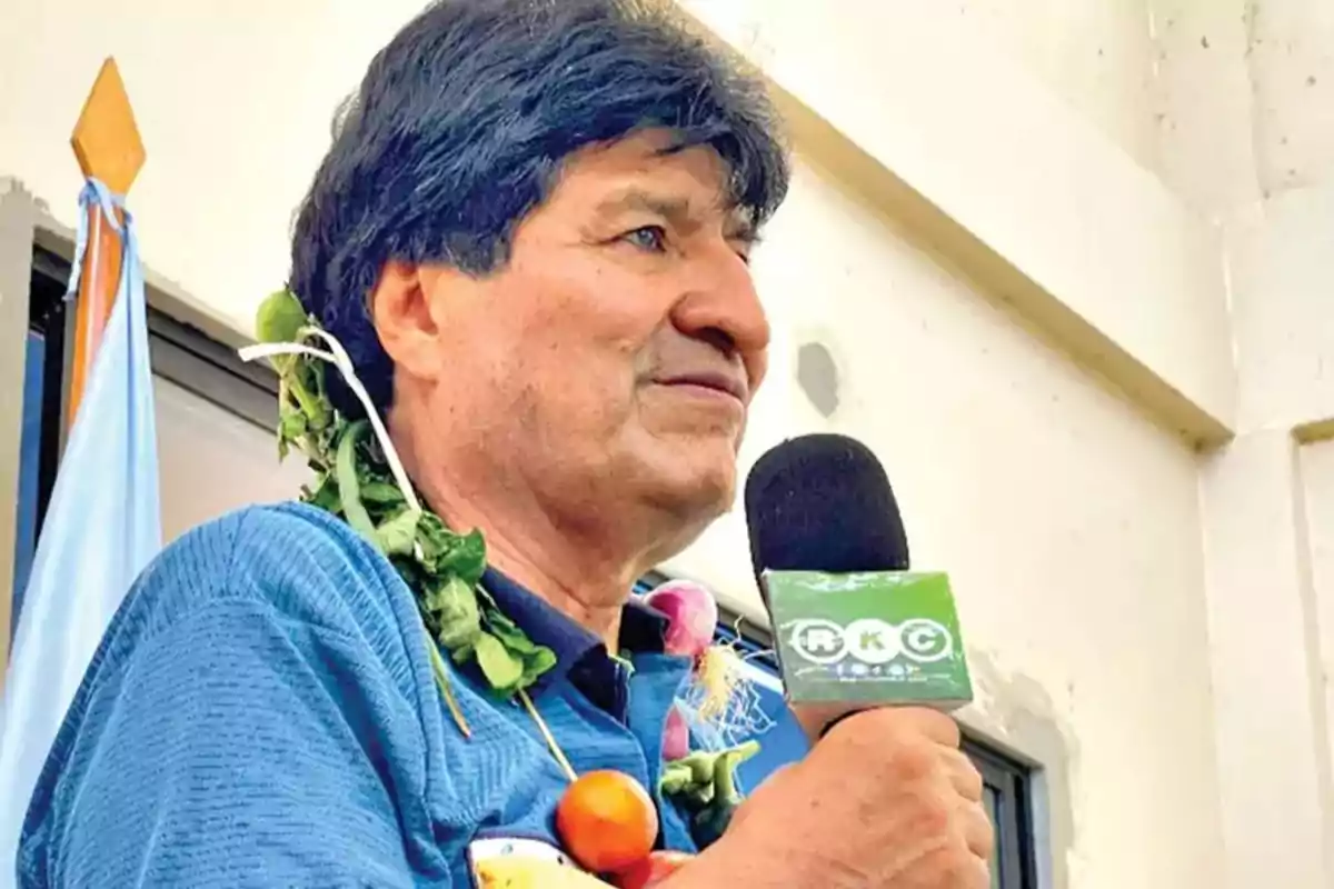 Un hombre con una guirnalda de vegetales alrededor del cuello sostiene un micrófono con un logotipo verde mientras está de pie frente a una bandera.