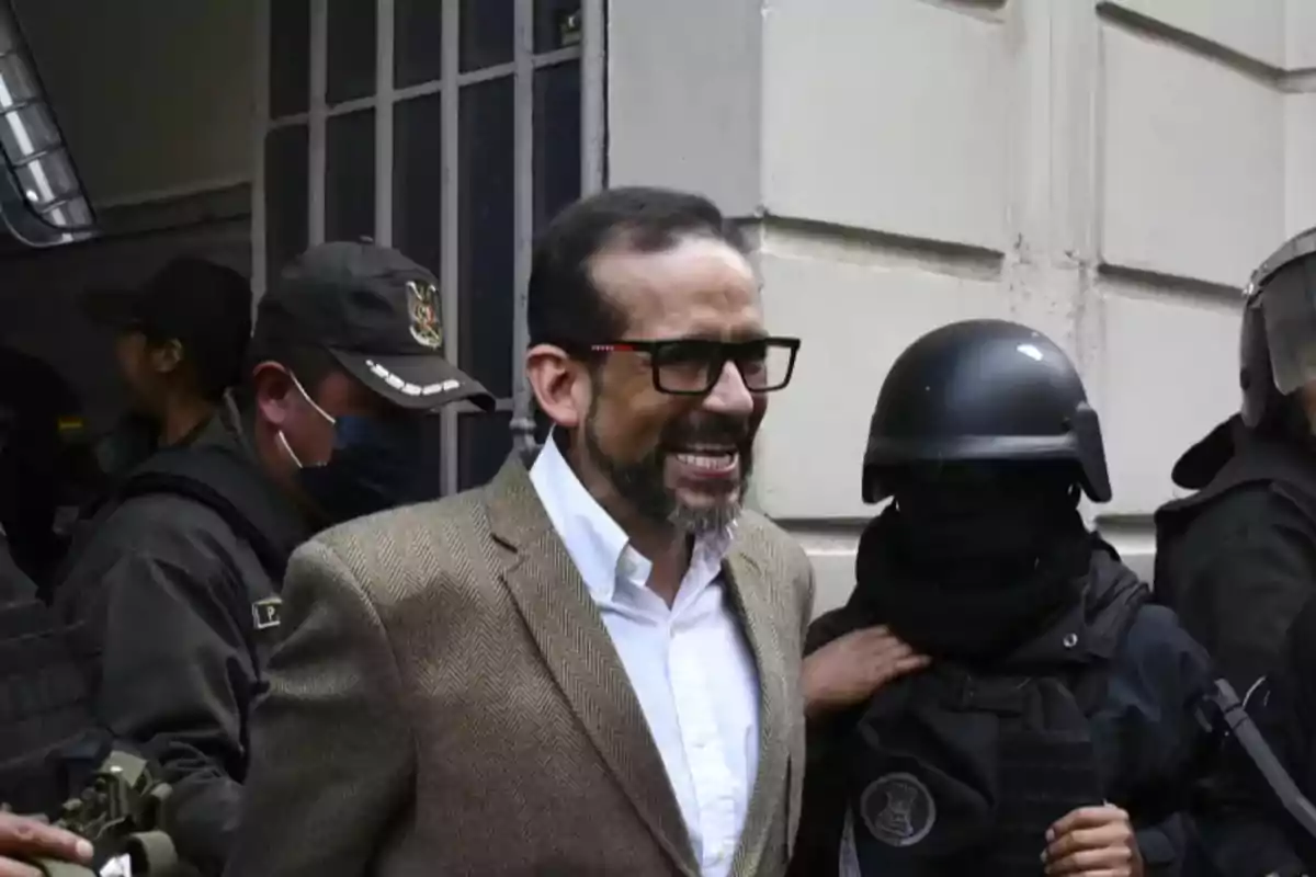 A man with glasses and a beard smiles while being escorted by uniformed police officers.