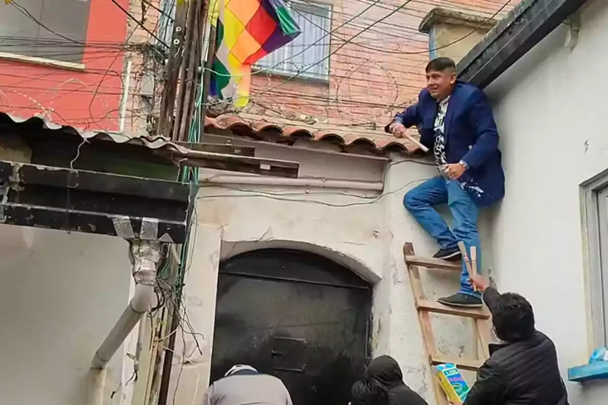 A man climbs a wooden ladder leaning against a wall while other people watch from below; in the background, a colorful flag and electrical wires can be seen.