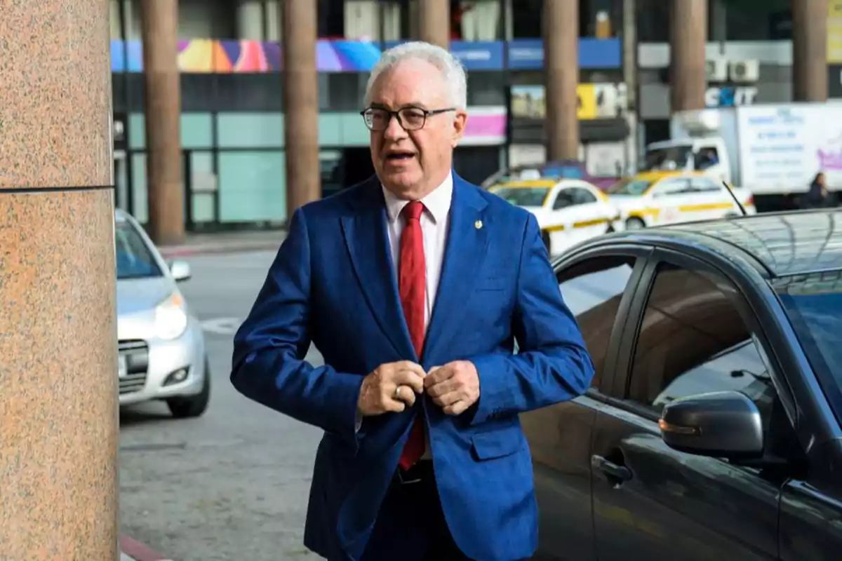 Gray-haired man with glasses and a blue suit buttoning his jacket next to a car on an urban street Gray-haired man with glasses and a blue suit buttoning his jacket next to a car on an urban street
