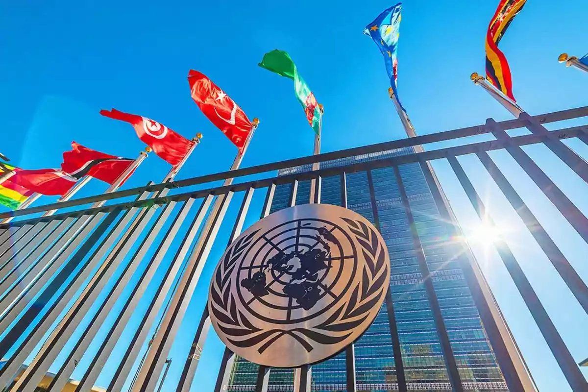 Edificio de las Naciones Unidas con banderas ondeando y el emblema de la ONU en una reja bajo un cielo azul. Edificio de las Naciones Unidas con banderas ondeando y el emblema de la ONU en una reja bajo un cielo azul.